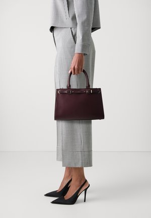 Femme en tailleur à carreaux tenant un sac à main en cuir bordeaux foncé, portant des escarpins noirs à bout pointu, debout devant un fond uni.