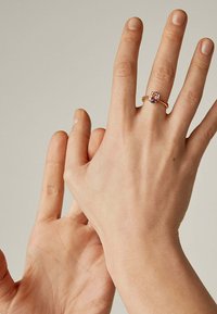 Anillo de oro con una piedra preciosa morada, con un diseño de banda retorcida. Manos mostradas con una mano extendiéndose hacia la otra.