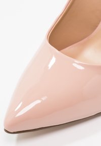 Chaussure à talon haut rose pâle brillant à bout pointu avec surface lisse et doublure intérieure beige sur fond blanc.