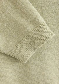 Lichtgroene sweaterstof van dichtbij, met een gladde textuur en geribbeld manchet-detail. Het materiaal lijkt zacht en comfortabel.