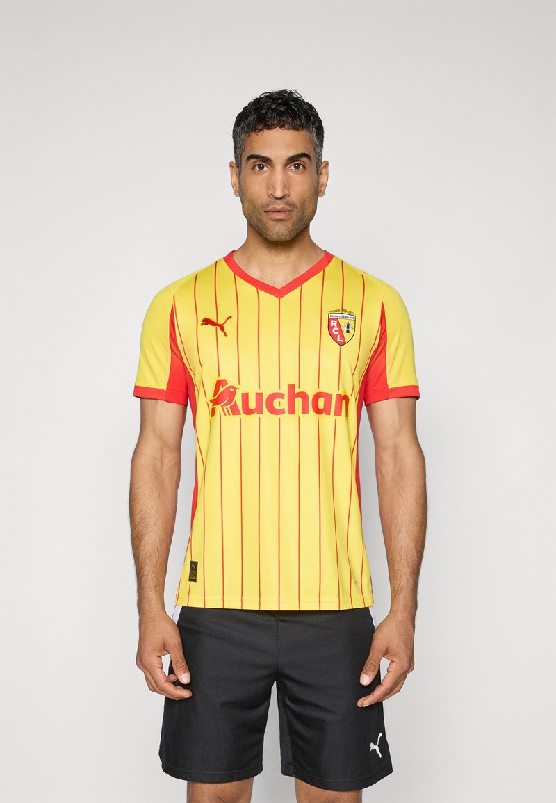 Un uomo è in piedi indossando una maglietta da calcio gialla brillante con strisce rosse, dotata di un logo, abbinata a pantaloni corti neri. Sfondo neutro.