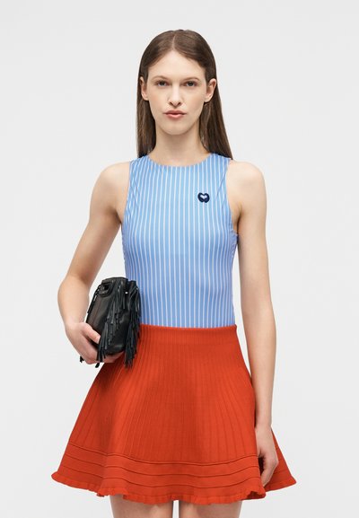 Femme portant un haut rayé bleu sans manches et une jupe plissée orange, tenant un sac à main noir à franges dans sa main gauche.