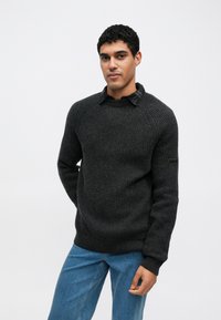 Pulover gri închis cu dungi, cu decolteu rotund și mâneci lungi, având o etichetă cu logo pe brațul stâng, asortat cu blugi albastru deschis.