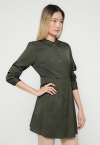 Žena s rovnými svetlo ombre vlasmi, oblečená v tmavozelenej košeľovej šatách s vyhrnutými rukávmi, stojaca s jednou rukou na bedrách.