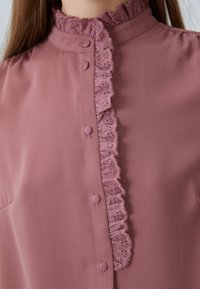 Blusa mauve con collo alto e merletti, chiusura frontale con bottoni e tessuto morbido testurizzato. Include bottoni decorativi lungo il davanti.