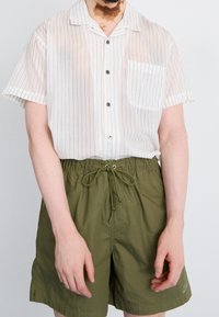 Muž v bílém krátkém rukávu s knoflíkovým zapínáním a tenkými černými pruhy, v olivově zelených šortkách na stažení, stojící na prostém pozadí.