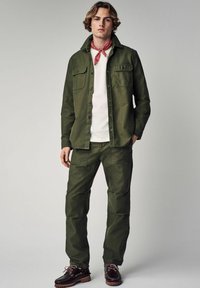 Olive green shirt jacket with two chest pockets, paired with matching pants. White t-shirt underneath, red bandana, and brown shoes.