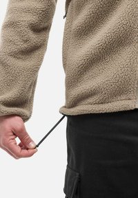 Main tenant un cordon de serrage noir au niveau de l'ourlet d'une veste en polaire beige portée sur un pantalon noir avec une poche latérale.