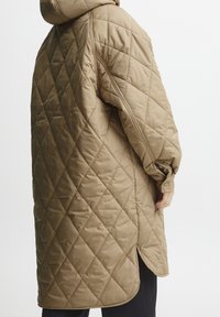 Vadderad, beige oversized jacka med huva, prydd med ett diamantmönster, sidofickor och detaljer vid ärmarna, tillverkad av ett smidigt, isolerat material.