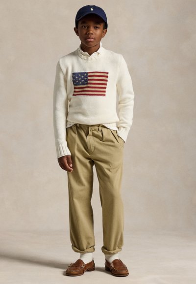 Poika, jolla on tummansininen lakki, valkoinen villapaita, jossa on amerikkalaisen lipun kuva, khakiväriset leikkerit housut, valkoiset sukat ja ruskeat loafersit, seisoo käsi taskussa.