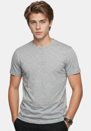 Jeune homme aux cheveux châtain clair portant un t-shirt gris uni à manches courtes et un pantalon noir, les mains dans les poches, fond neutre.
