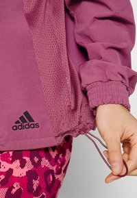 Chaqueta deportiva morada con paneles de malla, puños elásticos y dobladillo con cordón. Logo negro de Adidas en la parte inferior izquierda. Combinando con leggins rosas con estampado.