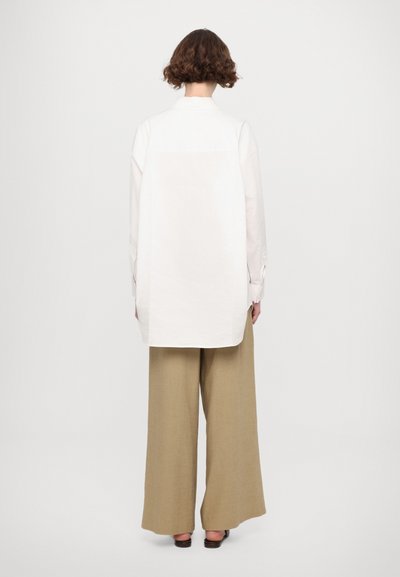 Camicia bianca oversize con retro lungo, abbinata a pantaloni beigi larghi. La camicia presenta un colletto e polsini con bottoni, con una texture di tessuto liscia.