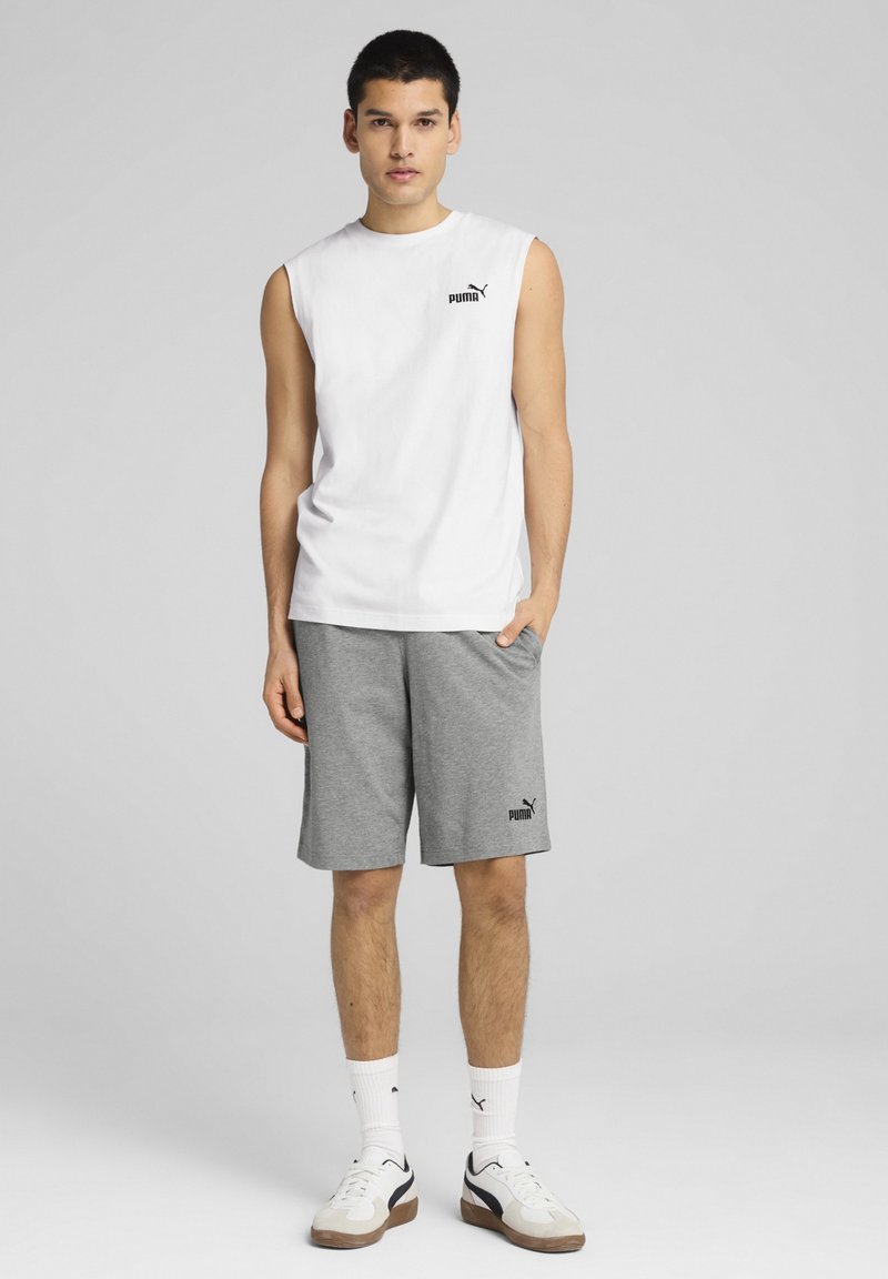 Balta bez piedurknēm krekls ar melnu Puma logotipu, salikts ar pelēkiem šortiem, kuriem arī ir logotips, nēsājot baltas zeķes un kedas ar melniem akcentiem.