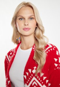 Roter Strickcardigan mit weißen geometrischen Mustern, getragen über einem weißen Hemd. Das Modell hat lange, wellige, blonde Haare und farbenfrohe, hängende Ohrringe.