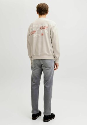 Osoba stoi tyłem, ubrana w jasnobeżową bluzę z napisem „Authentic New Edition 2023” oraz szare dżinsy na jednolitym tle.