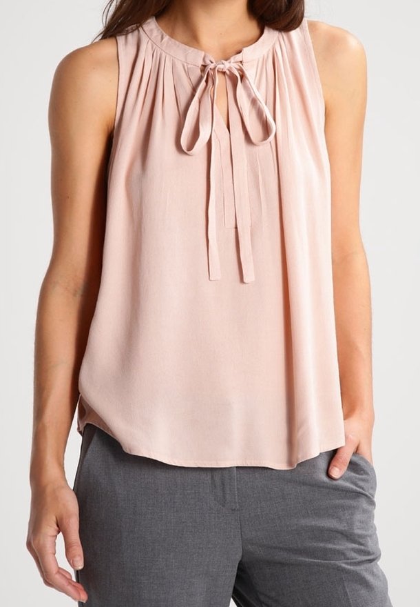 Blouse sans manches rose clair avec un lien au niveau du col, portée avec un pantalon gris, sur une personne debout avec une main dans la poche.