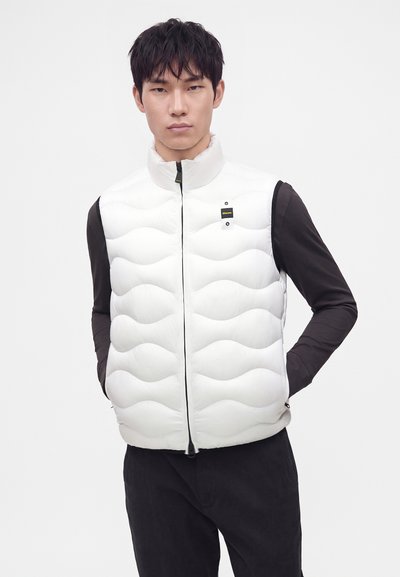 Hombre vestido con un chaleco blanco acolchado sobre una camisa negra de manga larga, de pie con las manos en los bolsillos del chaleco frente a un fondo liso.