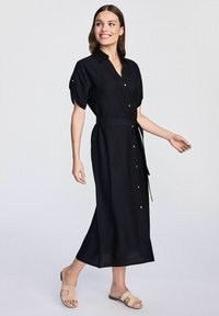 Czarna lniana sukienka na guziki z krótkimi bufkami, z paskiem w talii i bocznymi rozcięciami, zestawiona z jasnobeżowymi klapkami.