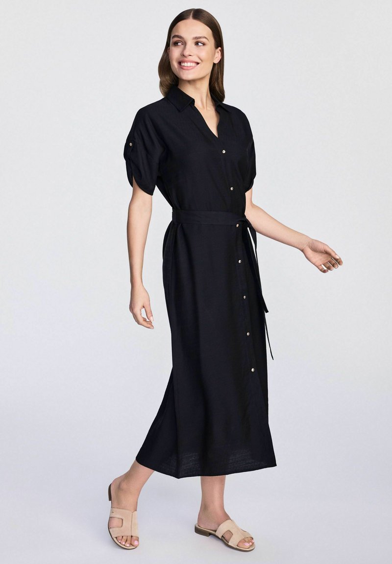 Czarna lniana sukienka na guziki z krótkimi bufkami, z paskiem w talii i bocznymi rozcięciami, zestawiona z jasnobeżowymi klapkami.