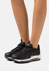 Fekete Nike Air Max 97 sportcipők többrétegű dizájnnal rendelkeznek, hálós és bőr felsőrésszel, fehér középtalppal és fekete gumitalppal.