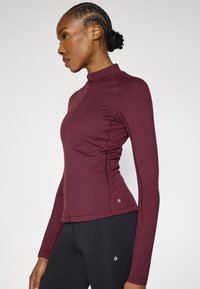 Tumši sarkans sportisks garroku tops ar rāvējslēdzēju, augstu apkakli, piegulošu dizainu, plakanām šuvēm un mazu logotipu apakšējā pusē. Kombinēts ar melnām legingām.