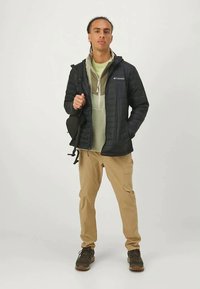 Μαύρο μπουφάν με επένδυση και φερμουάρ μπροστά, χακί cargo παντελόνια και ανοιχτό πράσινη κουκούλα. Άνδρας κρατάει μια μαύρη τσάντα ώμου. Καφέ ορειβατικά παπούτσια.