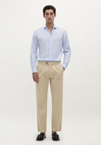 Camisa abotonada de color azul claro con mangas largas, combinada con pantalones beige de pinzas. Unos zapatos mocasines negros completan el conjunto.