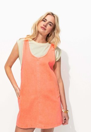 Robe en denim de couleur corail vif avec un décolleté rond, deux poches latérales et une coupe décontractée, superposée sur une chemise vert clair.