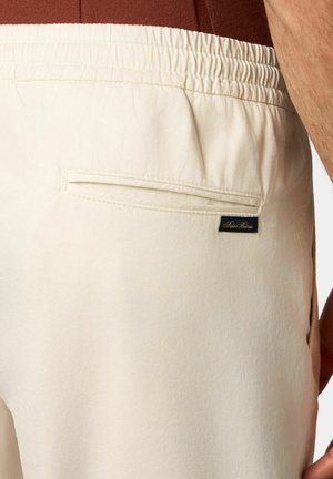 Beige stoffen shorts met een elastische tailleband, voorzien van een achterzak en een klein donker label met tekst aan de rechterkant.
