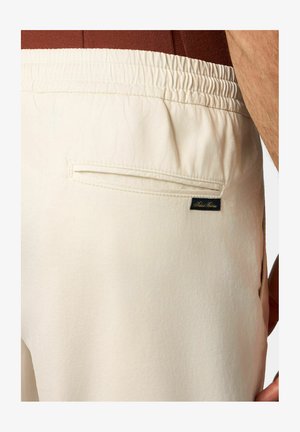 Shorts di tessuto beige con cintura elastica, dotati di una tasca posteriore e una piccola etichetta scura con testo sul lato destro.