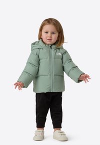 Light green puffer jacket with a hood, front zipper, and logo detail. Paired with black pants and light-colored shoes.