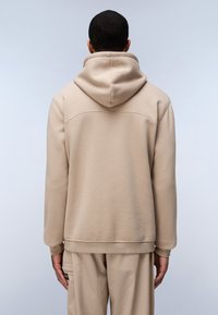 Sudadera con capucha de color beige, con un corte relajado, textura suave y un detalle de costura en la parte superior de la espalda. Combinada con pantalones beige a juego.