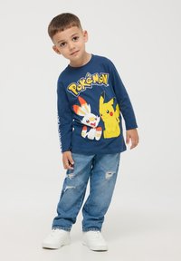 Granatowa koszulka z długim rękawem z grafikami Pikachu i Scorbunny, w połączeniu z jasnoniebieskimi przetartymi dżinsami i białymi trampkami.