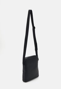 Bolso cruzado de cuero negro con un diseño plano, cierre de cremallera y una larga correa de tela. Textura suave, forma minimalista.