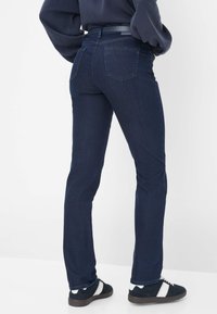 Persoon in donkerblauwe jeans, zwarte riem, marineblauwe trui en zwarte sneakers met witte strepen, staande op een witte vloer tegen een witte achtergrond.