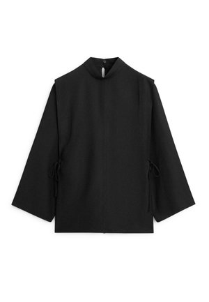 Czarna bluzka z długim rękawem, wysokim kołnierzem, zapięciem na guziki z tyłu przy szyi oraz wiązaniami po bokach na jednolitym białym tle.