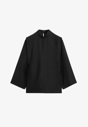 Blusa nera a maniche lunghe con collo alto, chiusura con bottoni sul retro del collo e dettagli con fiocchi laterali su sfondo bianco uniforme.