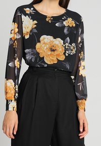 Langärmlige Bluse aus transparenter schwarzer Stoff mit großen orangen und cremefarbenen Blumenmustern. Manschettendetails mit gemusterten Akzenten.