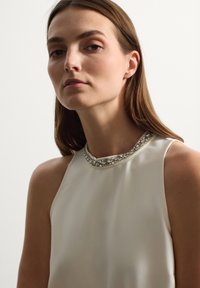 Top sin mangas de color crema con un cuello alto, que presenta un collar decorativo de cuentas. Tejido suave y liso con un brillo sutil.