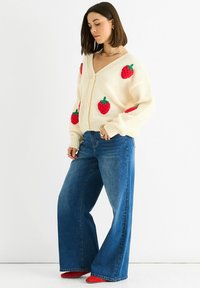 Romige gebreide cardigan met rode aarbeienapplicaties, V-hals en knoopsluiting, in combinatie met lichtblauwe uitlopende jeans en rode schoenen.