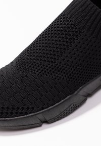 Gros plan d’une basket slip-on noire en maille montrant une tige texturée et respirante ainsi qu'une semelle épaisse texturée sur fond blanc.