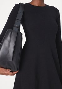 Femme portant une robe moulante noire à manches longues avec un logo en relief sur la poitrine, tenant un sac à bandoulière en cuir noir avec une large sangle.