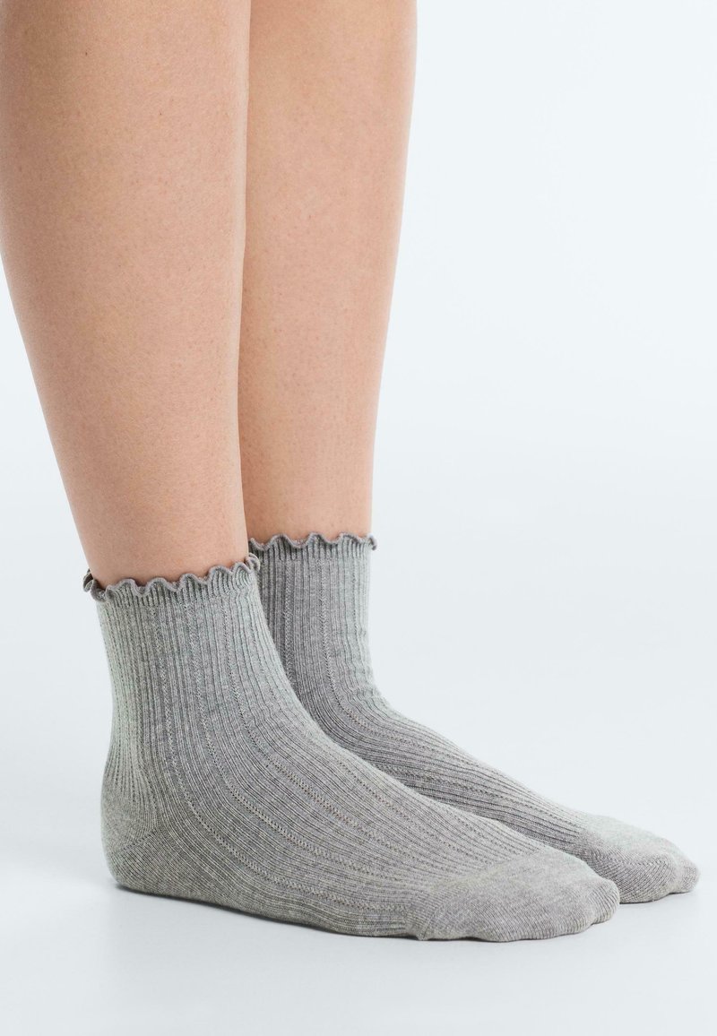 Jambes portant des chaussettes courtes côtelées gris clair avec des bords festonnés sur un fond clair uni.