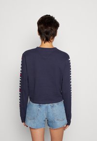 Námořnický dlouhý rukáv s kulatým výstřihem, se červeným a bílým textem podél rukávů. V kombinaci se světlemodrými džínovými šortkami.