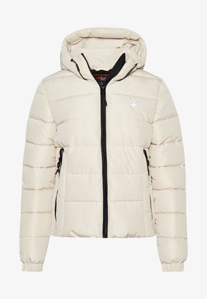 Veste matelassée crème légèrement rembourrée avec une fermeture éclair noire, poches latérales zippées, une capuche et des poignets élastiques. Présente un logo sur la poitrine.