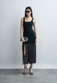 Γυναίκα φοράει ένα μαύρο αμάνικο φόρεμα με ασημένια τρουκς και σκισίματα, μαύρα ψηλοτάκουνα σανδάλια, μαύρα γυαλιά ηλίου και κρατάει ένα ασημένιο τσαντάκι.