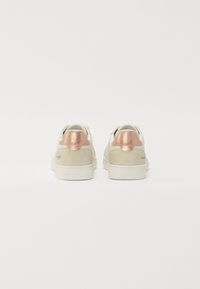 Et par hvite lavtopp sneakers med beige semsket hæler og metallisk roségull detaljer på en enkel hvit bakgrunn.