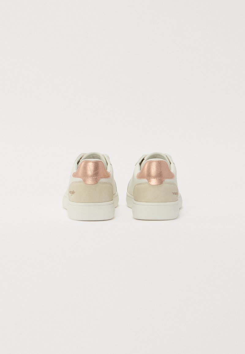 Un par de zapatillas deportivas bajas blancas con talones de ante beige y acentos metálicos en oro rosa sobre un fondo blanco liso.