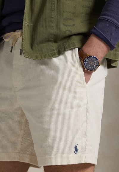 Κρεμ σορτς από κορντόνι με ανάγλυφη υφή και διακριτικό ναυτικό λογότυπο στο τελείωμα· συνδυασμένα με πράσινο μπουφάν και καφέ ρολόι.